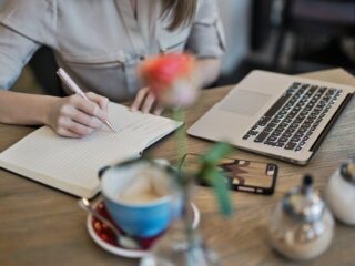 Free Woman writing in a notebook with a laptop and coffee cup on a desk. Ideal for workspace inspiration. Stock Photo