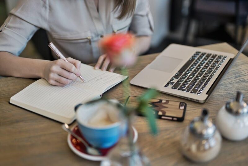 Free Woman writing in a notebook with a laptop and coffee cup on a desk. Ideal for workspace inspiration. Stock Photo