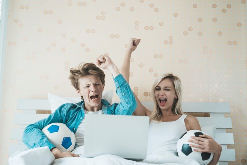 Happy couple watching a soccer match in bed while placing bets online